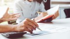 Two people sat at a table, one is writing in a book and the other is pointing a pen at a document